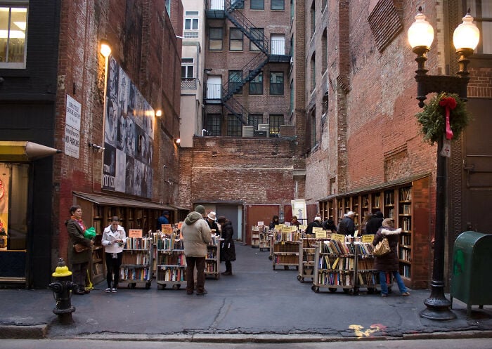 Livraria aconchegante ao ar livre num beco urbano com pessoas a consultar livros, rodeado por paredes de tijolo e candeeiros de rua antigos.