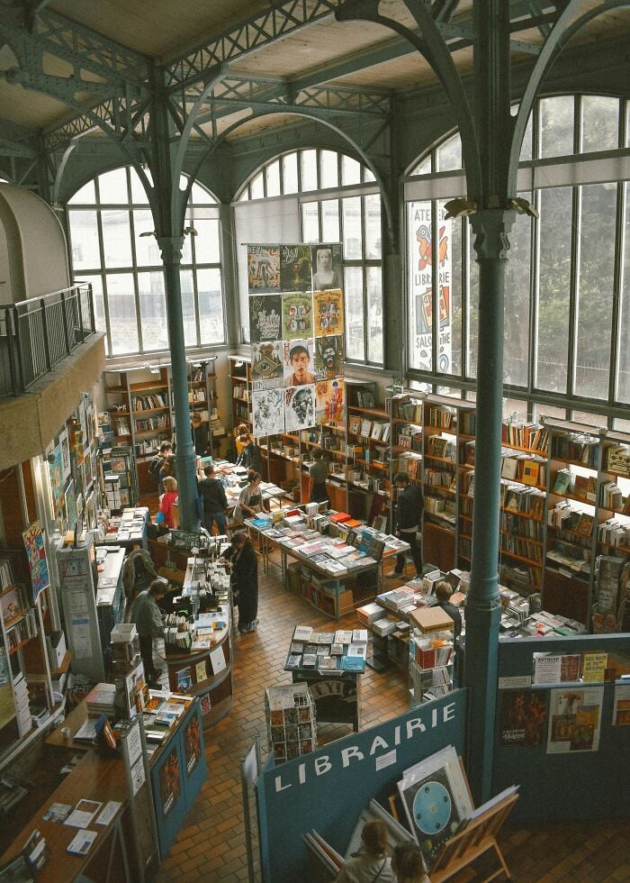 Biblioteca espaçosa e grandiosa com grandes janelas arqueadas, altas colunas de metal e prateleiras cheias de livros e visitantes navegando.