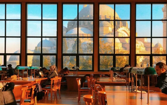 Interior aconchegante de biblioteca com grandes janelas mostrando arquitetura grandiosa e pessoas lendo e estudando em mesas de madeira.