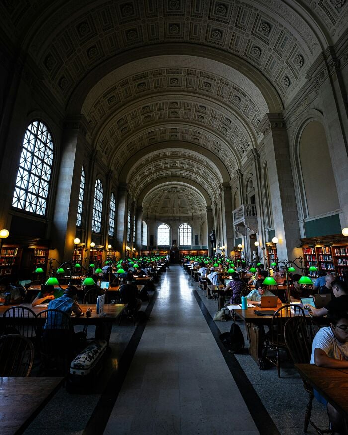 Interior grandioso de biblioteca com tetos altos em arco, grandes janelas e fileiras de mesas de estudo com luminárias verdes e leitores.