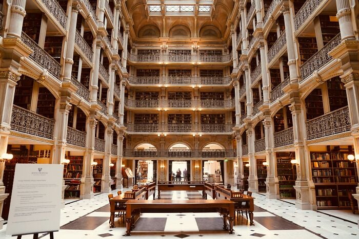 Interior grandioso de biblioteca com vários níveis de estantes, colunas ornamentadas e uma ampla área de leitura com mesas e cadeiras de madeira.