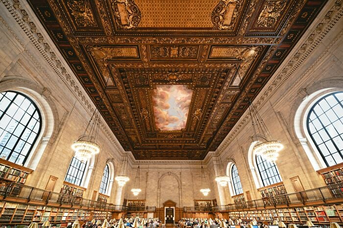 Interior grandioso de biblioteca com teto ornamentado, grandes janelas em arco, lustres e fileiras de estantes cheias de livros.